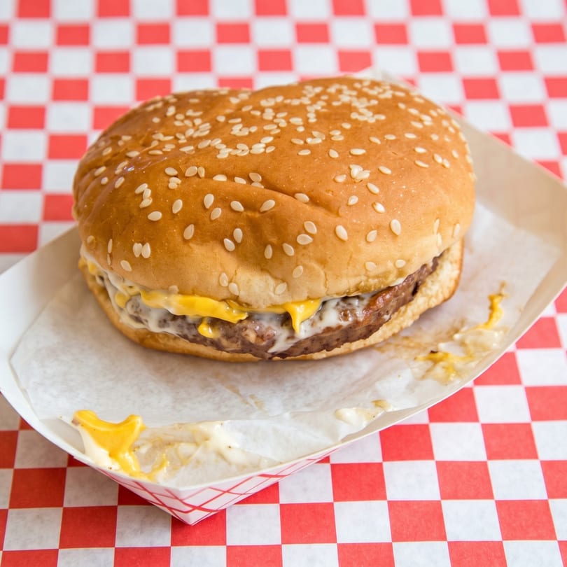 Cheeseburger with melted cheese and sauce in a paper tray on a red and white checkered tablecloth.
