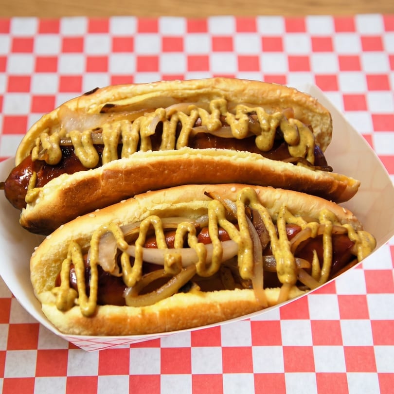 Two hot dogs with mustard and onions in a paper tray on a red and white checkered surface.